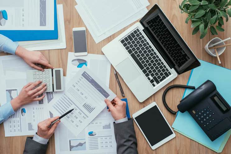 desk filled with paper documents, calculators, laptop, tablet and hands pointing to reports